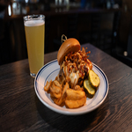 A plate with a sandwich topped with fried onions and cheese, served with waffle fries and pickles, next to a glass of light beer on a dark wooden table.