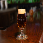 A glass of dark beer with a foamy head sits on a coaster on a wooden bar counter.