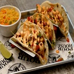 Three tacos with diced toppings and creamy sauce are served on a tray with a side of rice, a lime wedge, and branded paper underneath.