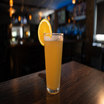 A tall glass of orange-colored beverage garnished with an orange slice sits on a wooden table in a dimly lit bar.