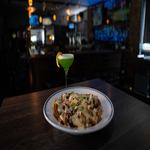 A bowl of loaded nachos topped with cheese and herbs sits on a table next to a green cocktail in a dimly lit bar.